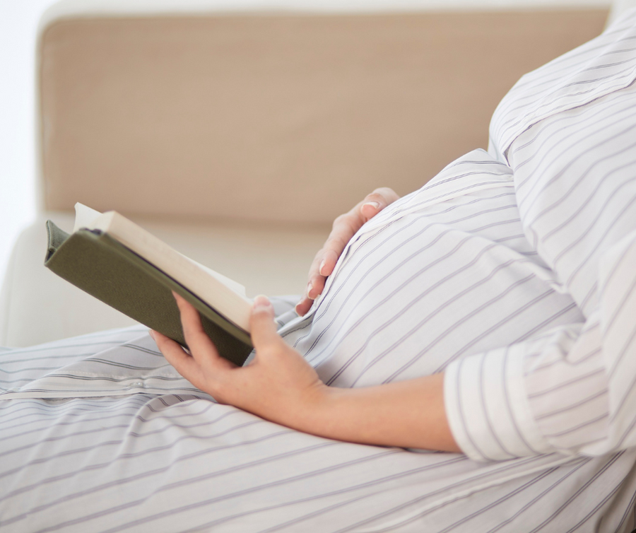 Schwangere Frau liest Buch und streichelt sich dabei den Bauch